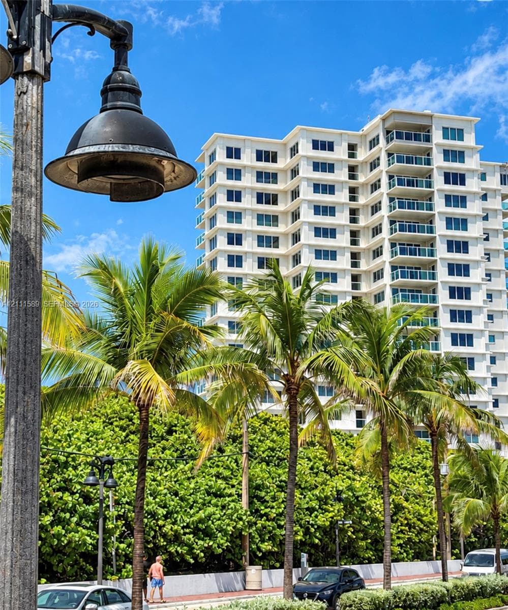 High-rise building featuring a white concrete exterior with multi-level balconies and uniform window placement, set against mature palm trees and custom black metal street lighting