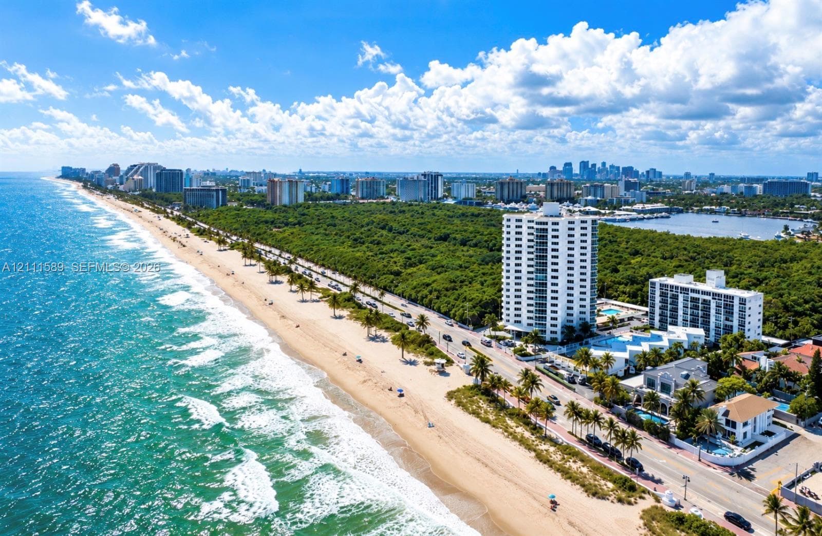 Coastal aerial perspective featuring direct oceanfront access, sandy shoreline, multi-story residential structures, lush tropical vegetation, and distant cityscape