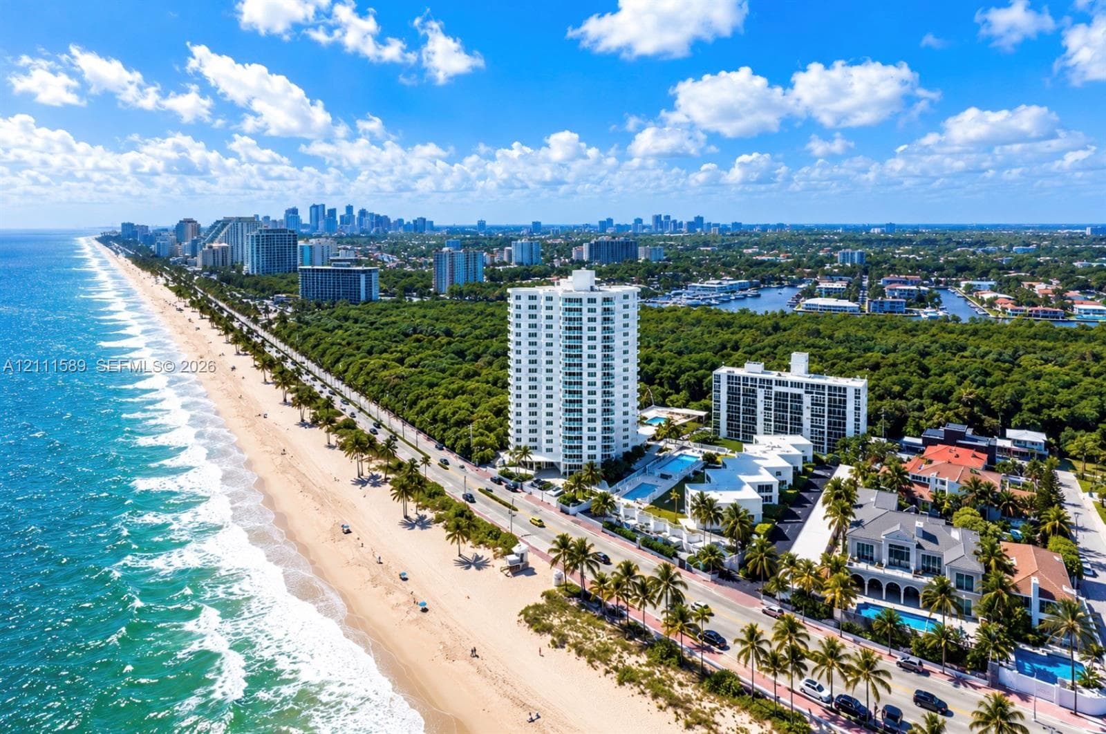 High-rise building featuring white exterior, balconies, and ground-level pool situated along a sandy coastline