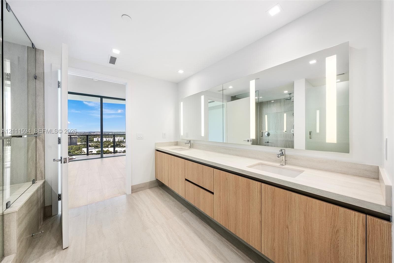 Floating vanity with wood-finish cabinetry, dual under-mount sinks, expansive wall-to-wall integrated mirror with vertical task lighting, and glass-enclosed shower area