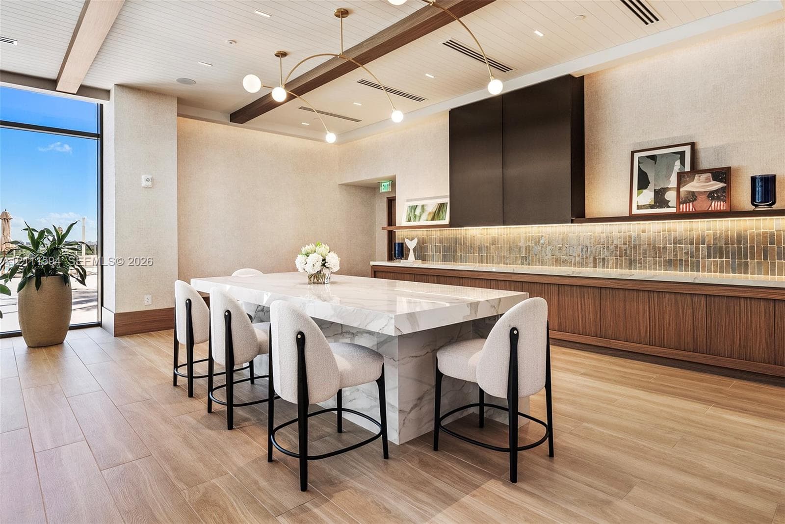 Waterfall-edge stone island with bar seating, custom wood-finish cabinetry, linear tile backsplash, wood-finish flooring, and recessed ceiling beams