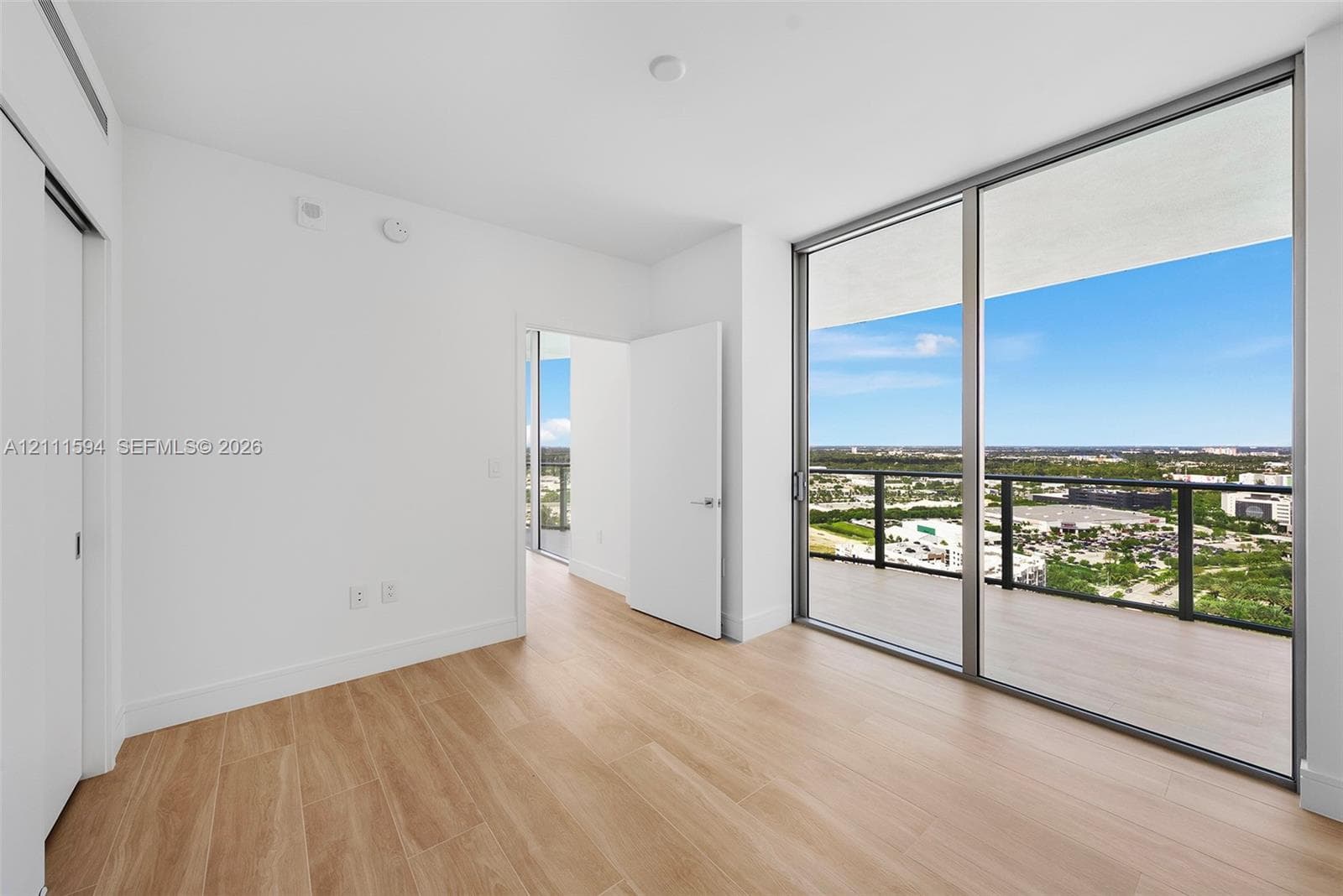 Floor-to-ceiling glass windows, private balcony with metal railing, wood-finish flooring, and crisp white wall finishes