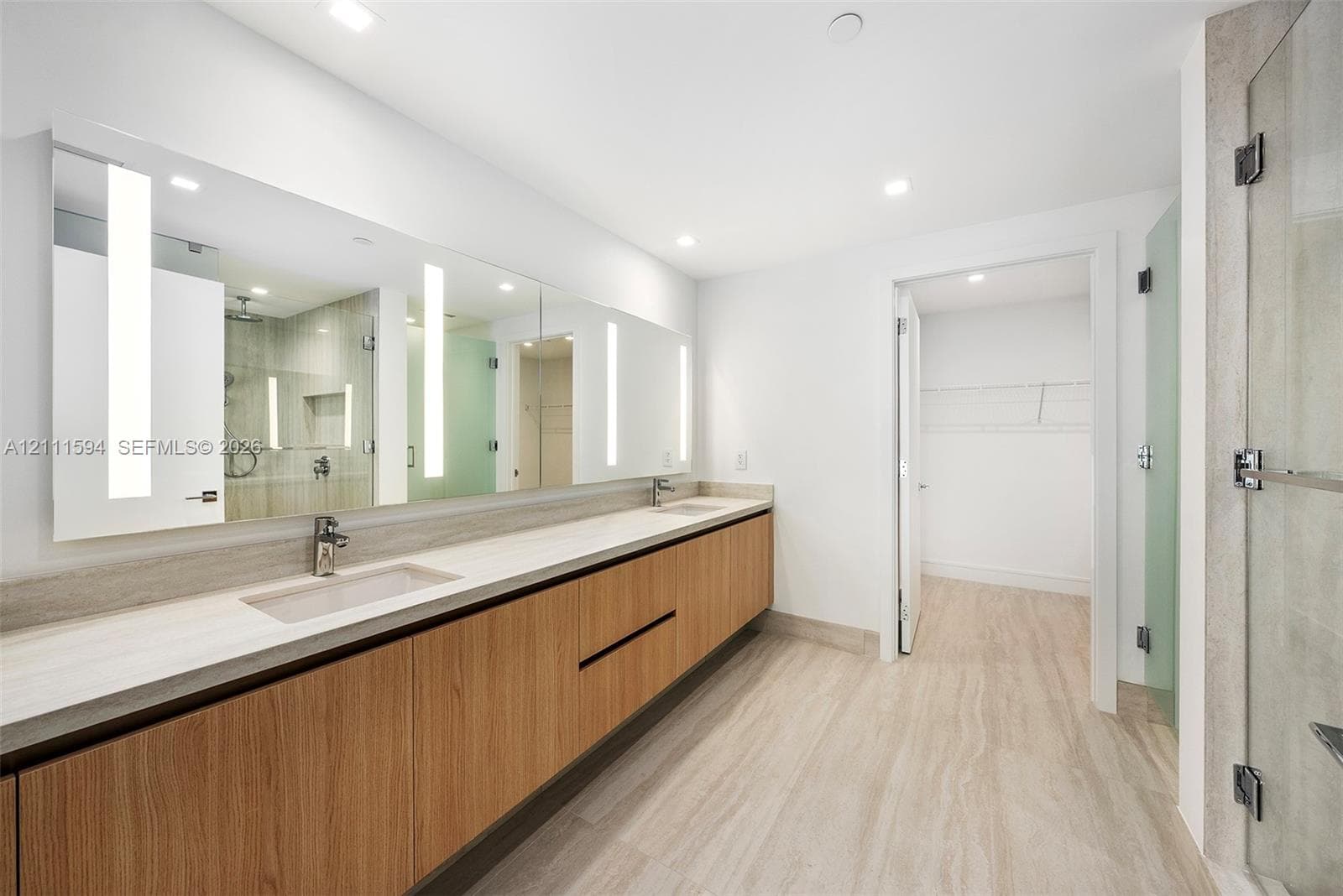 Floating wood-finish vanity with dual integrated sinks, expansive wall-to-wall mirror with integrated LED lighting, stone-finish flooring, and glass-enclosed shower