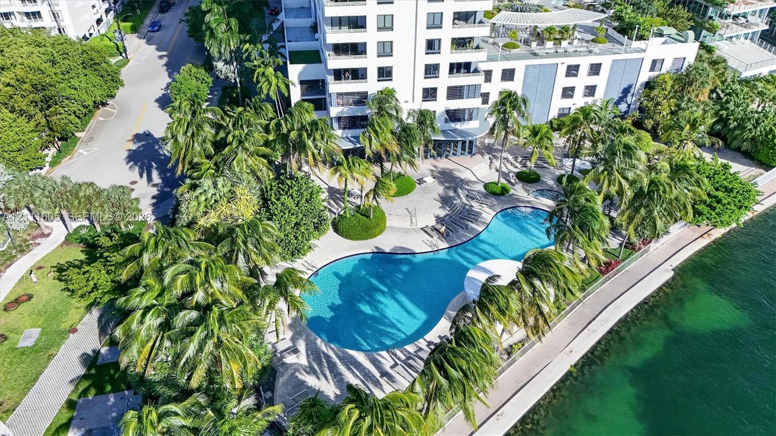 Multistory residential building with a curvilinear swimming pool, concrete hardscaping, mature palm trees, and direct waterfront access