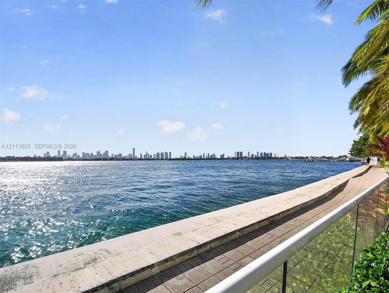 Waterfront property featuring a concrete seawall, paver-stone walkway, glass-panel railing, and tropical palm foliage
