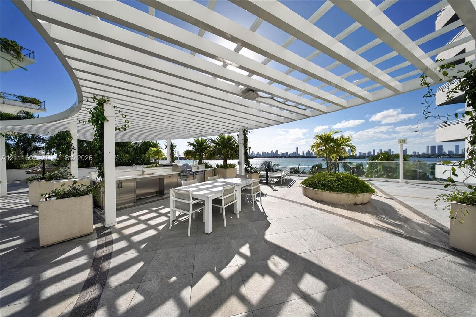 Expansive rooftop terrace featuring a curved white pergola, built-in stainless steel outdoor kitchen, stone tile flooring, and integrated concrete planters
