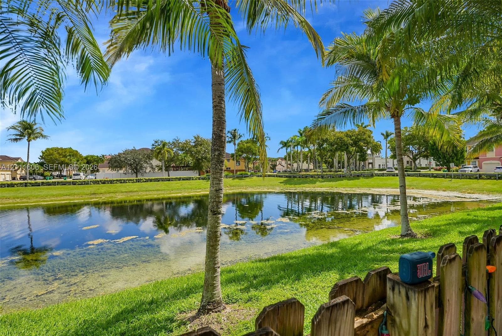 Waterfront lot featuring a pond, manicured lawn, and mature palm trees enclosed by a wooden picket fence
