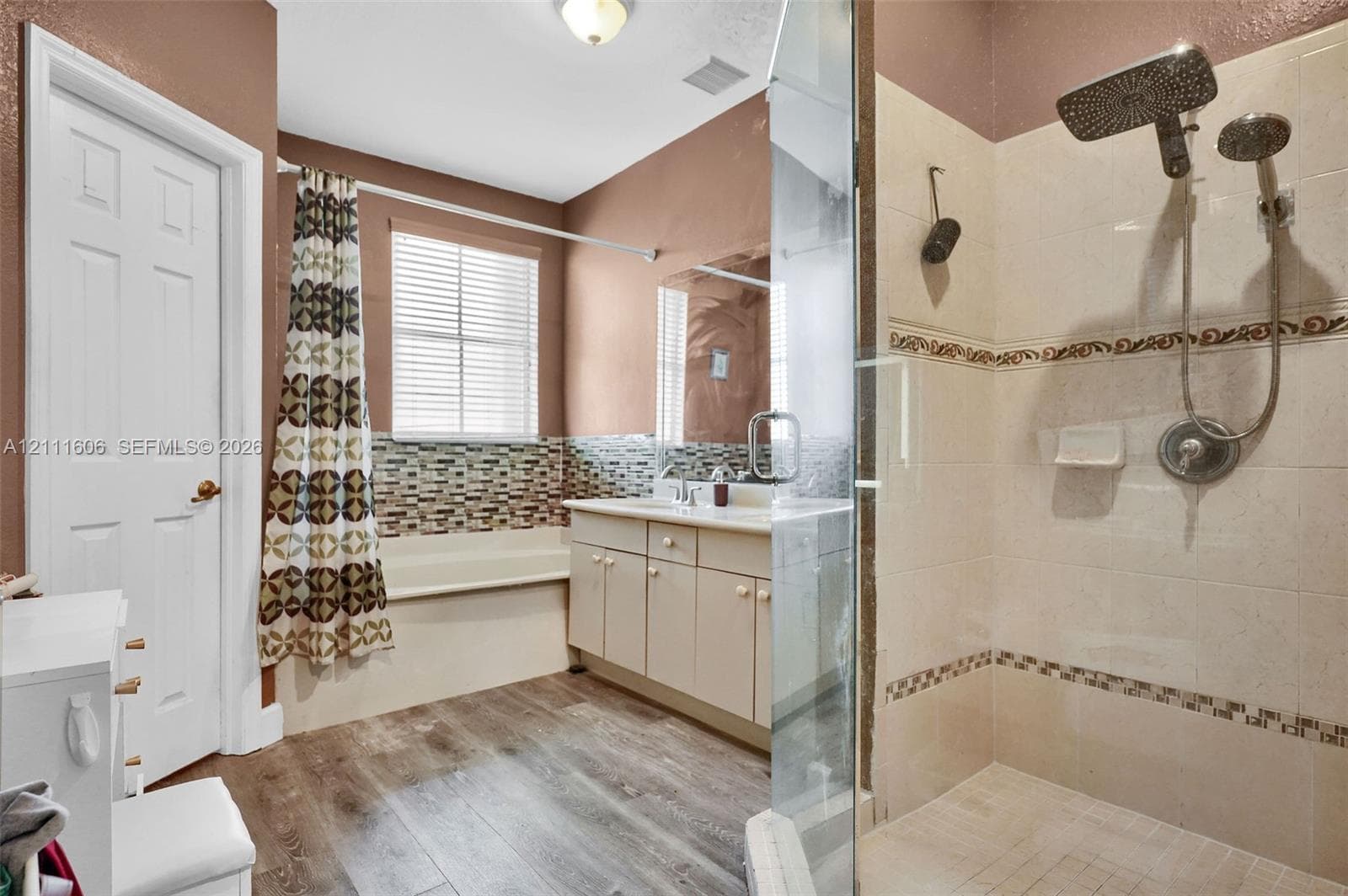 Bathroom featuring wood-finish flooring, a glass-enclosed shower with a rainfall showerhead, a built-in tub with decorative tile backsplash, and a vanity with a mirror