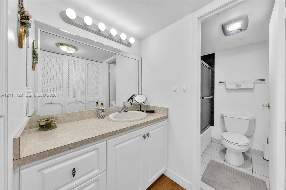 Bathroom featuring white cabinetry, solid-surface countertop with an integrated basin, large wall-mounted mirror, overhead multi-bulb lighting, and a tub-shower enclosure with tiled surround