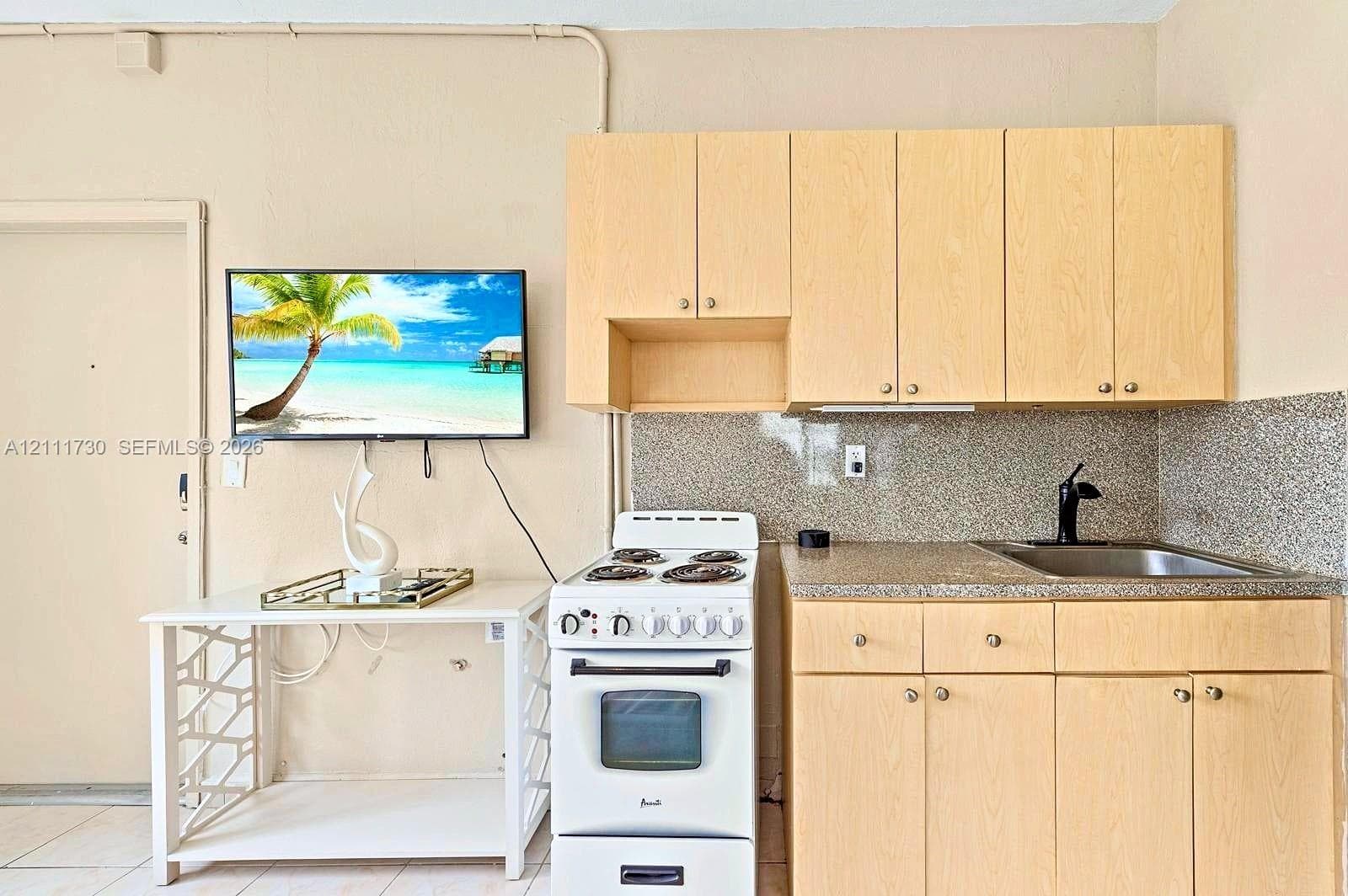 Kitchenette featuring light wood-finish cabinetry, speckled solid-surface countertops, integrated stainless steel sink with a matte black faucet, and a white freestanding electric range