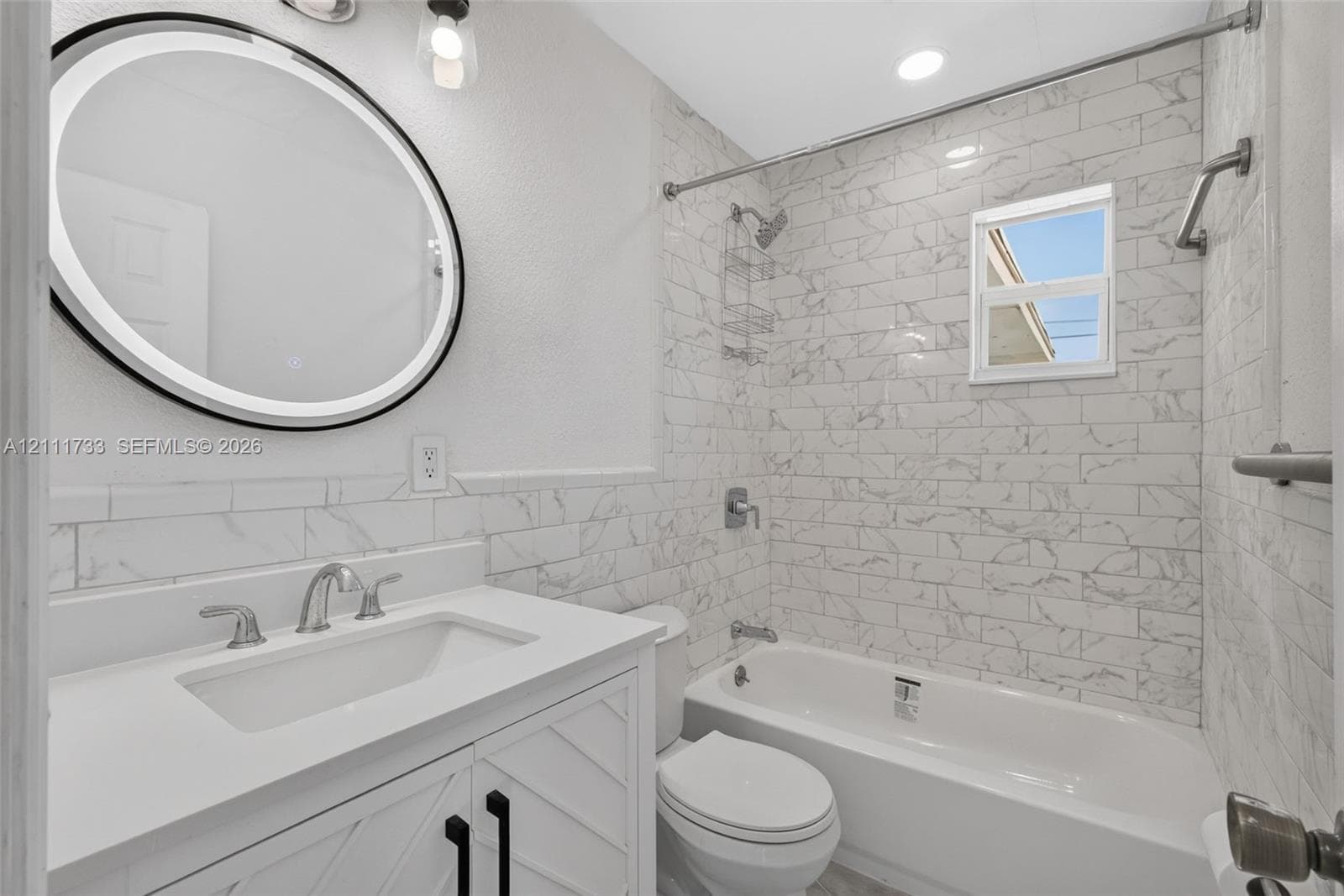 Main Bathroom featuring a white vanity with geometric cabinet detailing, a quartz countertop, an integrated vessel sink, a circular LED-backlit mirror, and white subway-patterned tile wall surrounds with a marble-style finish