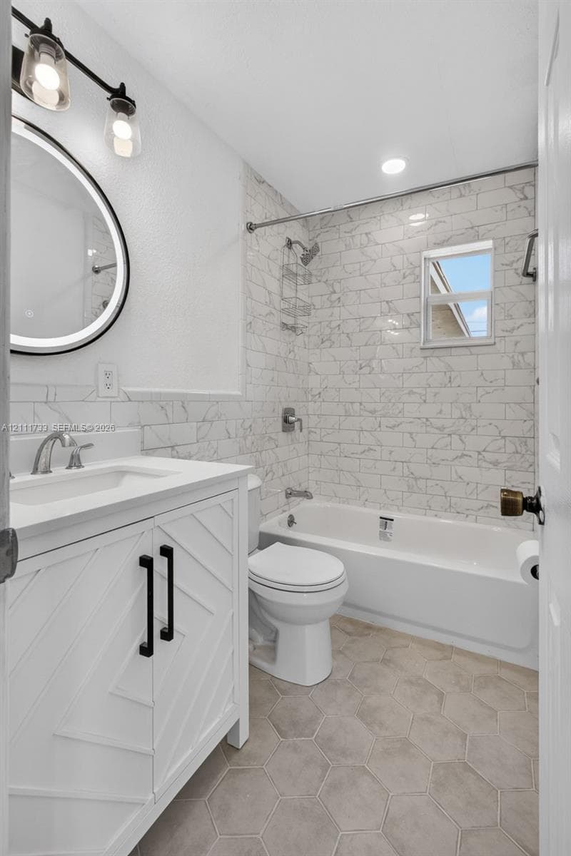 Main Bathroom with white vanity with chevron detailing and matte black hardware, illuminated circular vanity mirror, light-toned hexagonal floor tile, and subway-tiled tub surround