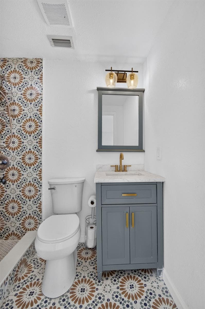 Ensuite Bathroom featuring patterned tile shower surround, grey vanity countertop, undermount sink, metallic faucet, and a wall-mounted light fixture