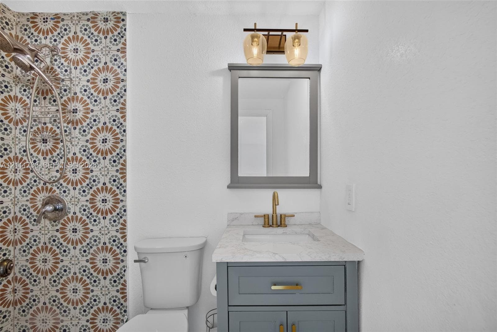 Ensuite Bathroom featuring patterned tile shower surround, grey vanity countertop, undermount sink, metallic faucet, and a wall-mounted light fixture.