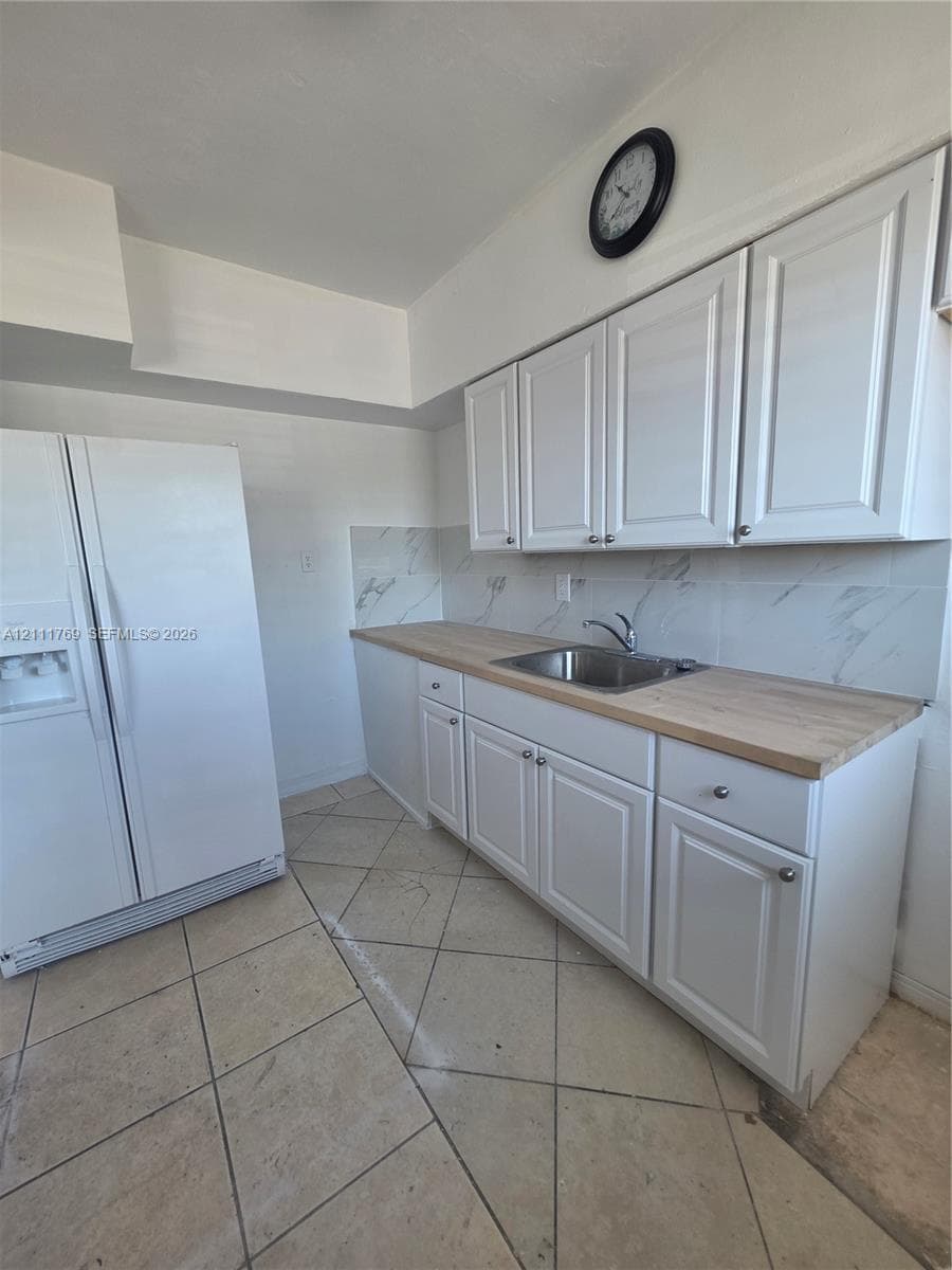 White cabinetry with stainless steel hardware, wood-finish countertop, stainless steel sink, stone-finish tile backsplash, and light-tone floor tiling