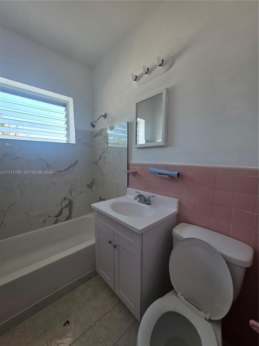 White vanity with integrated sink, porcelain bathtub surround featuring large-format stone-patterned tile, pink ceramic wall tile, and jalousie window