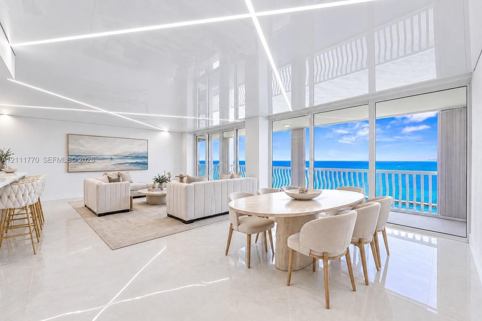 High-gloss reflective ceiling with integrated linear LED lighting, polished stone flooring, and expansive floor-to-ceiling glass sliding doors opening to an oceanfront balcony with structural railing