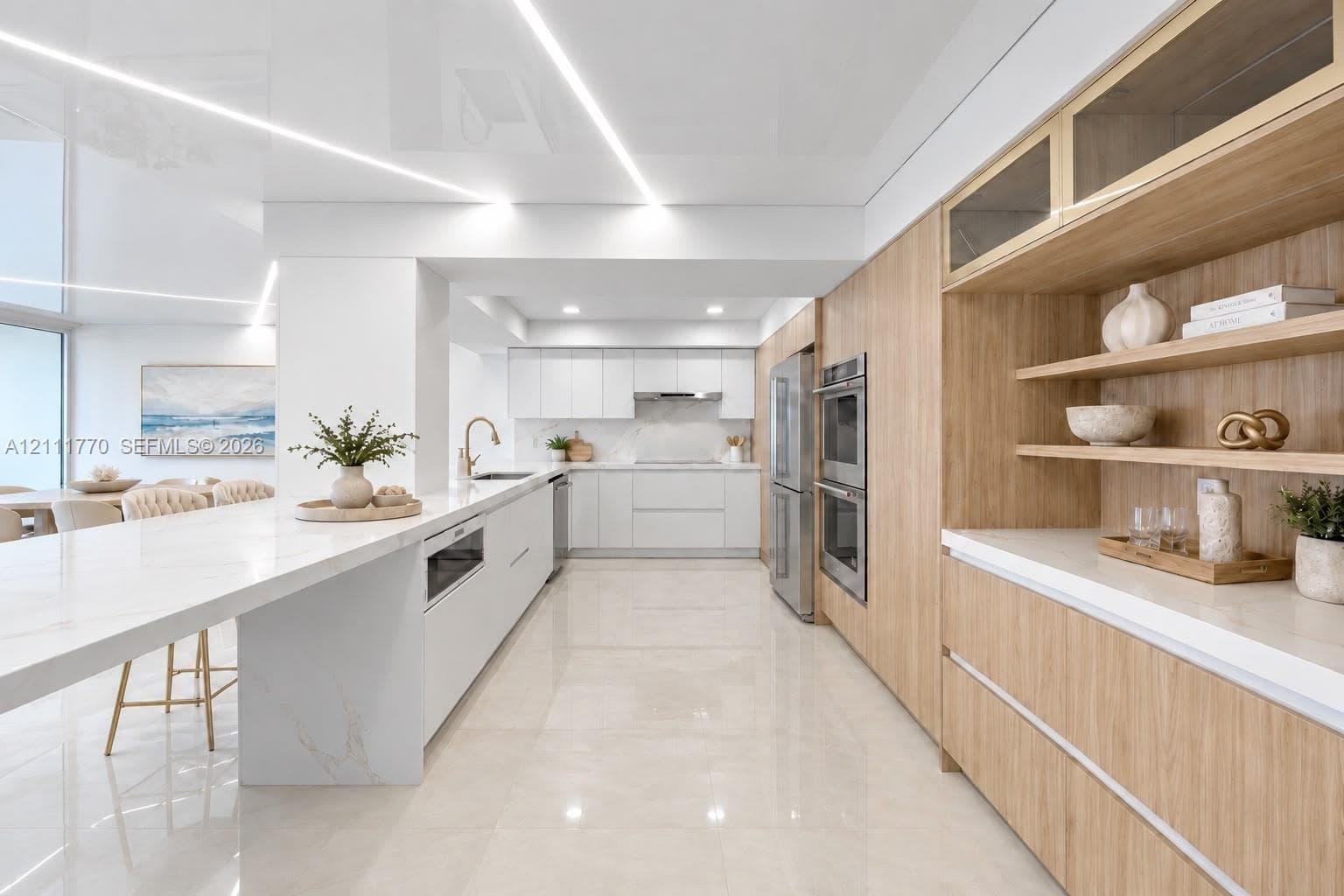 Contemporary kitchen featuring a sprawling waterfall-edge island, high-gloss flat-panel cabinetry, light-toned wood-finish cabinetry accents, integrated linear ceiling lighting, and stainless steel double wall ovens