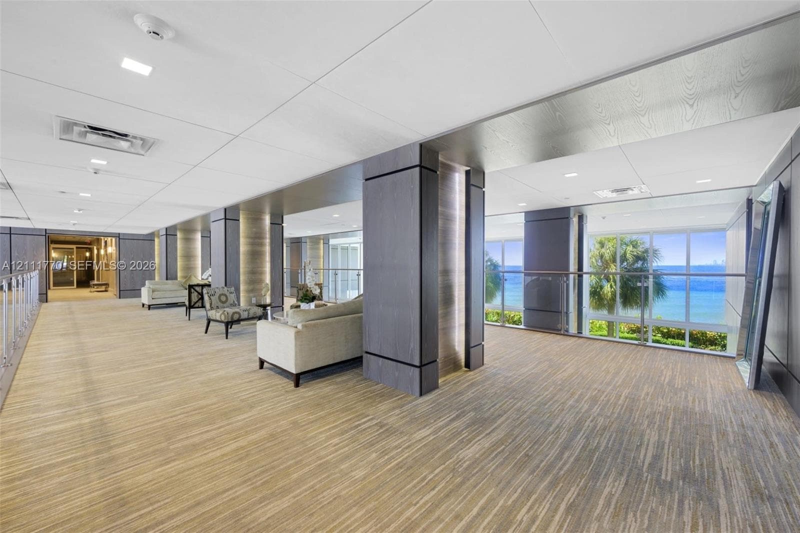 Expansive common area featuring wood-finish flooring, floor-to-ceiling windows with ocean scenery, dark wood-tone architectural columns, and recessed lighting