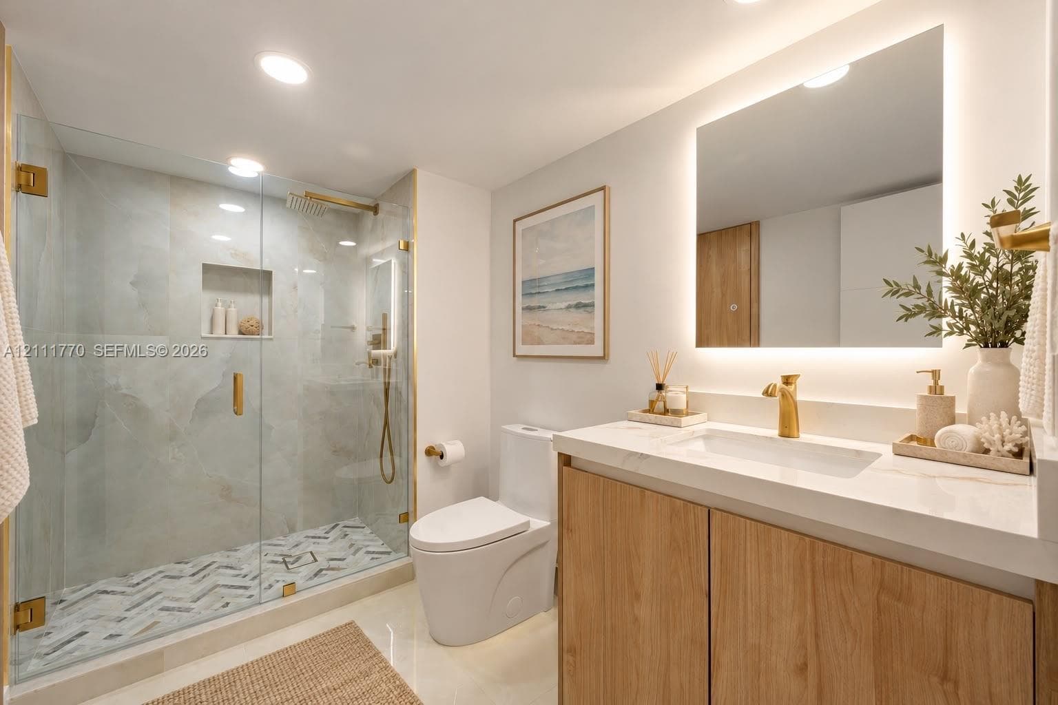 Bathroom featuring a frameless glass walk-in shower with stone tile surround, chevron-patterned floor tile, wood-finish vanity with a solid surface countertop, and a back-lit wall mirror
