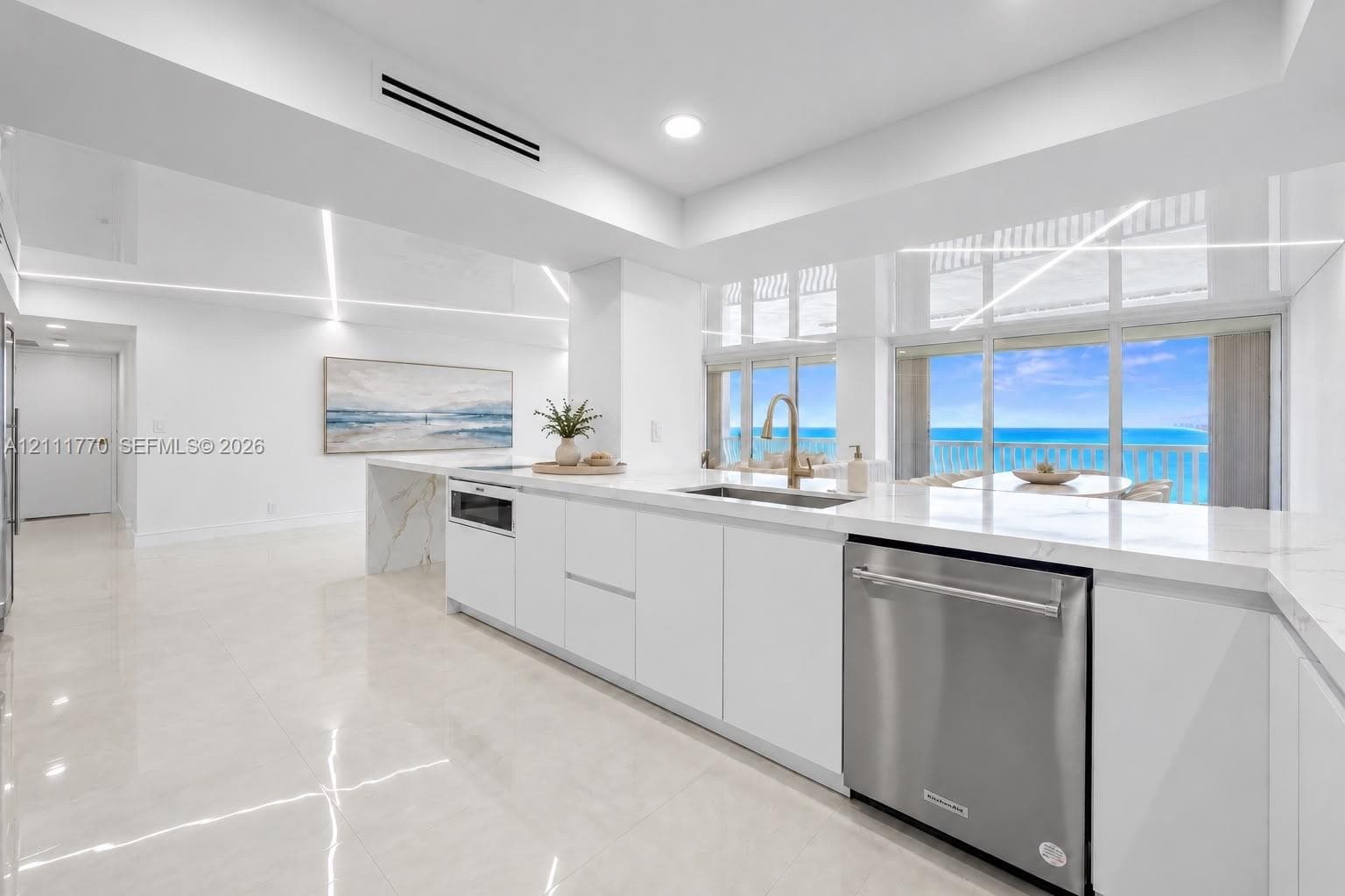Modern kitchen featuring sleek white cabinetry, stone-finish countertops, stainless steel appliances, and large-format tile flooring, complete with expansive floor-to-ceiling windows and ocean horizon scenery