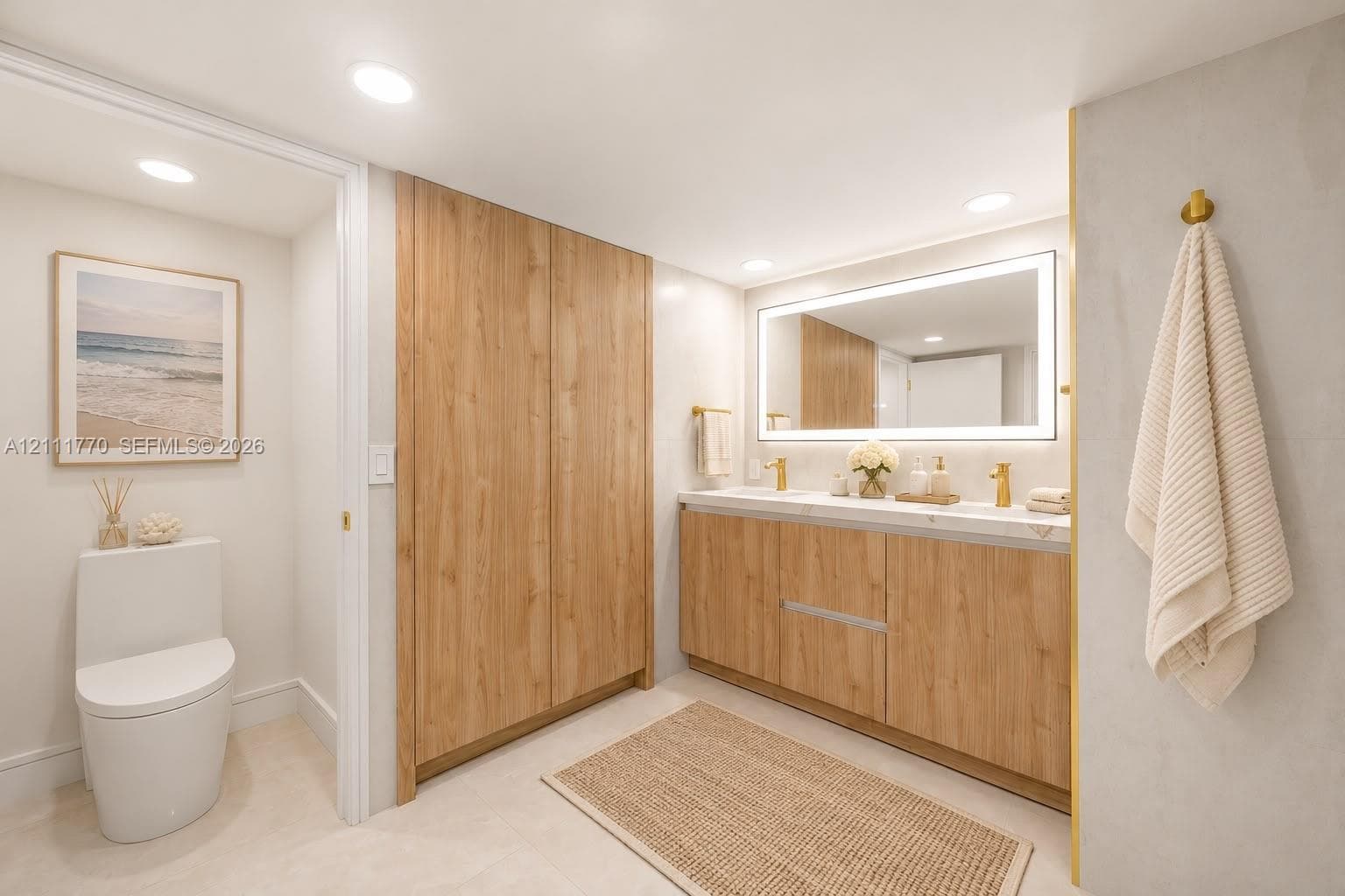 Bathroom featuring a wood-finish double vanity with dual gold-toned faucets, integrated solid-surface countertop, full-width illuminated vanity mirror, and built-in floor-to-ceiling cabinetry