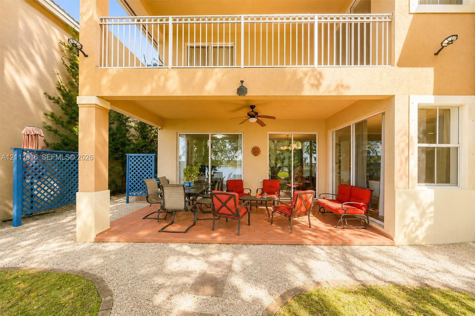 Covered ground-level patio with tiled flooring, upper-level balcony featuring white metal railings, integrated ceiling fan, and decorative blue privacy screens