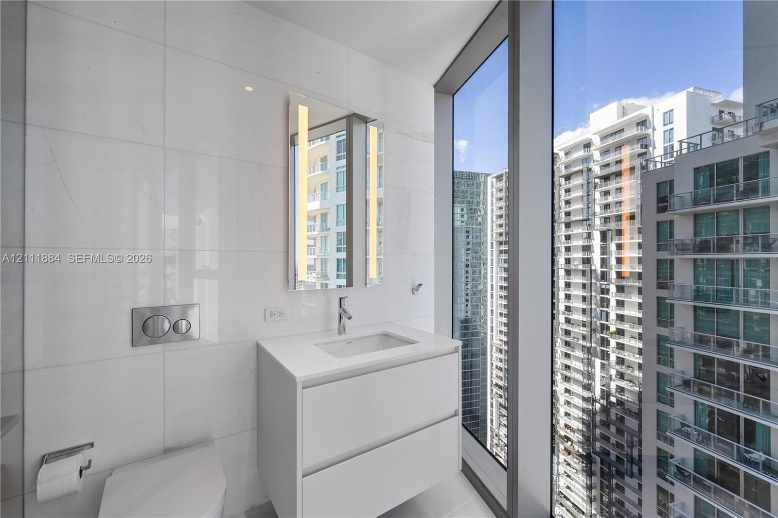Modern floating vanity with a white countertop and rectangular undermount sink, floor-to-ceiling windows, wall-mounted dual-flush toilet, and vertical LED-integrated mirror