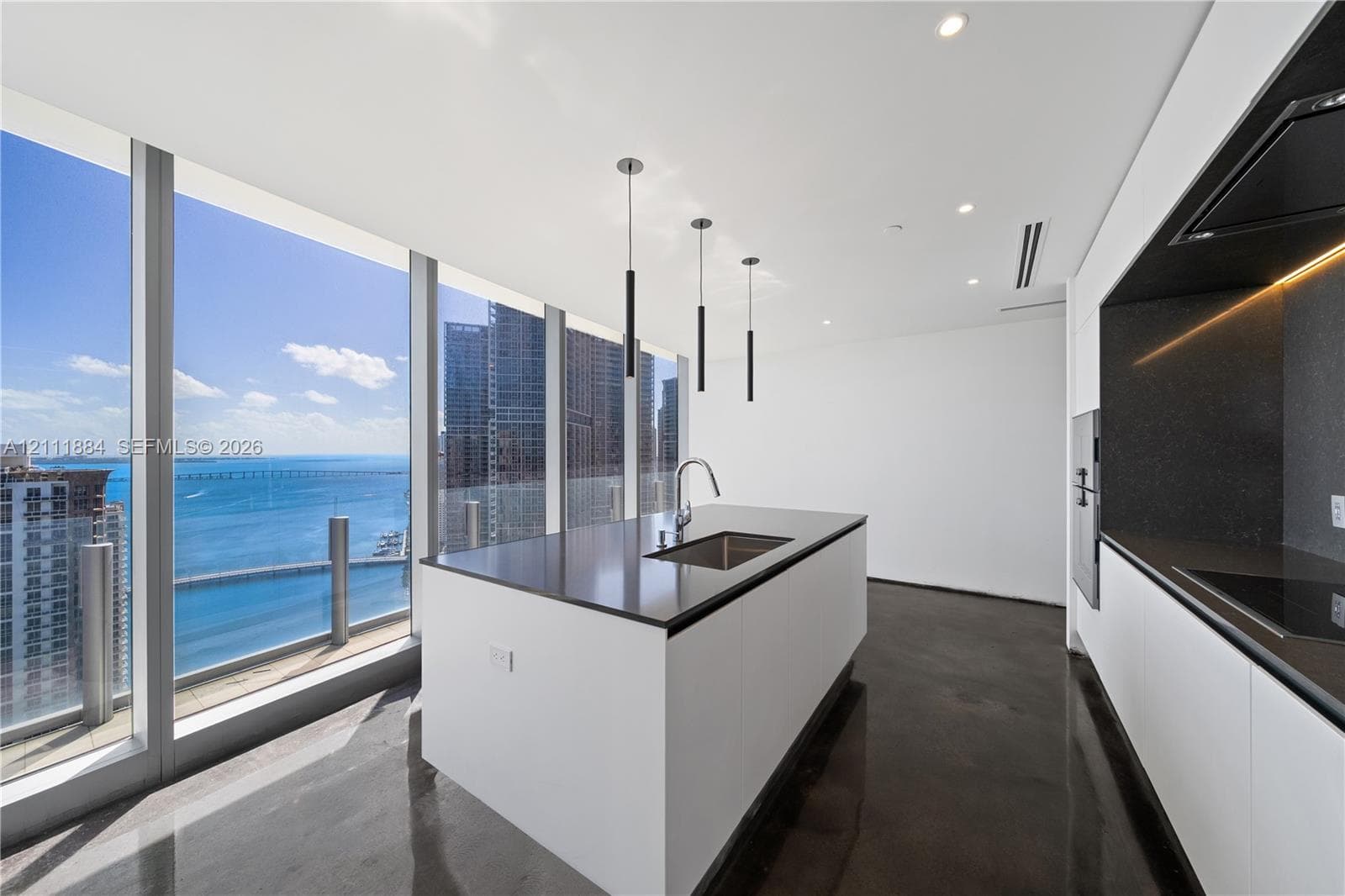 Kitchen featuring floor-to-ceiling windows, central island with an integrated stainless steel sink, polished concrete flooring, minimalist white cabinetry, and matte black pendant lighting