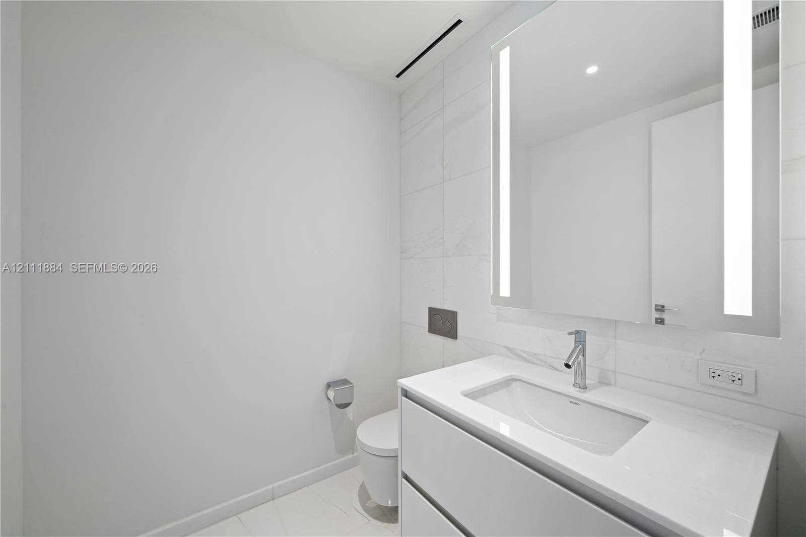 Floating vanity featuring a white countertop and rectangular basin, integrated LED-backlit mirror, wall-mounted faucet, and large-format tile wall