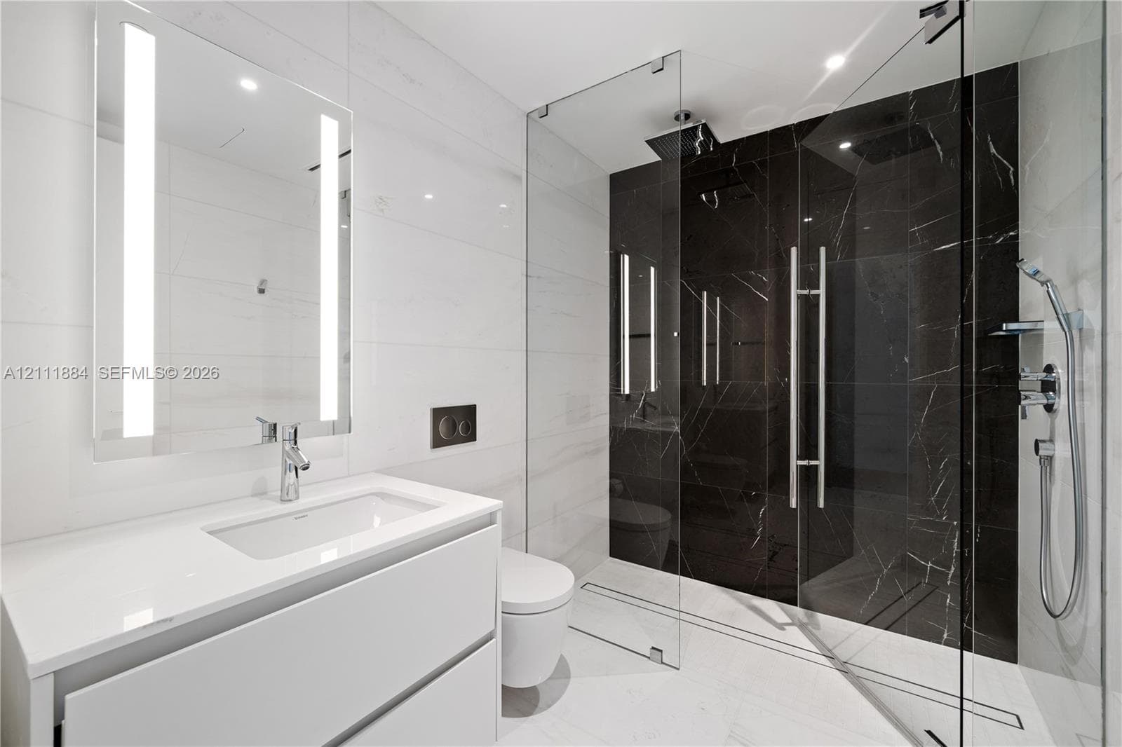 Floating vanity with a white countertop and rectangular undermount sink, integrated LED-lit mirror, wall-mounted toilet with dual-flush actuator, and glass-enclosed shower with contrasting large-format tile surrounds and rainfall showerhead