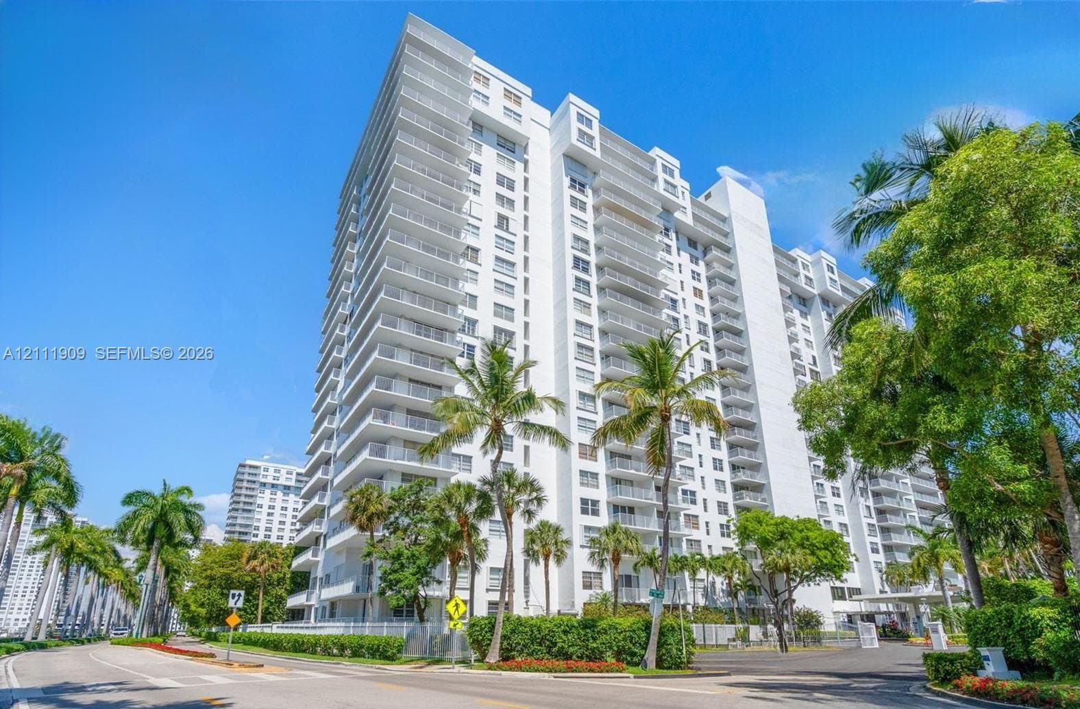 High-rise residential building featuring a white facade, wraparound balconies, glass railings, and manicured tropical landscaping