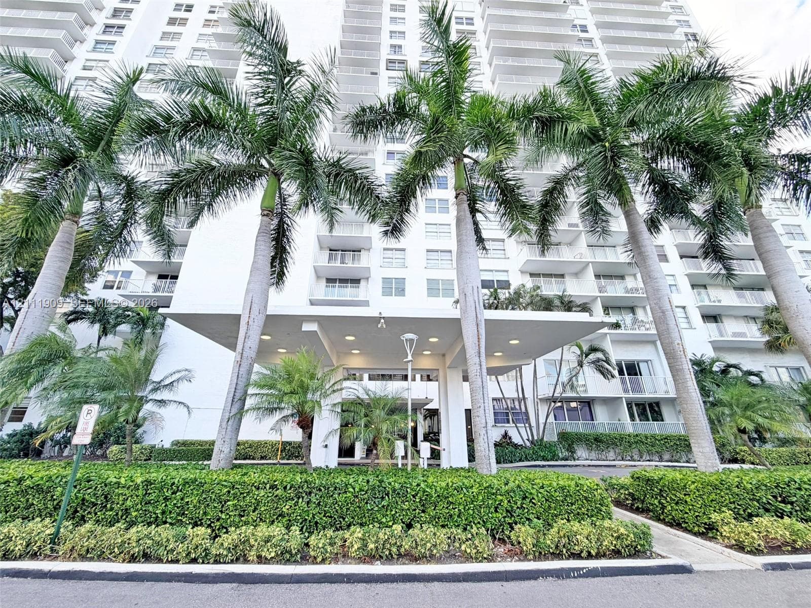 High-rise building exterior featuring a covered porte-cochere entry, wrap-around private balconies, recessed lighting, and manicured landscaping with mature palm trees