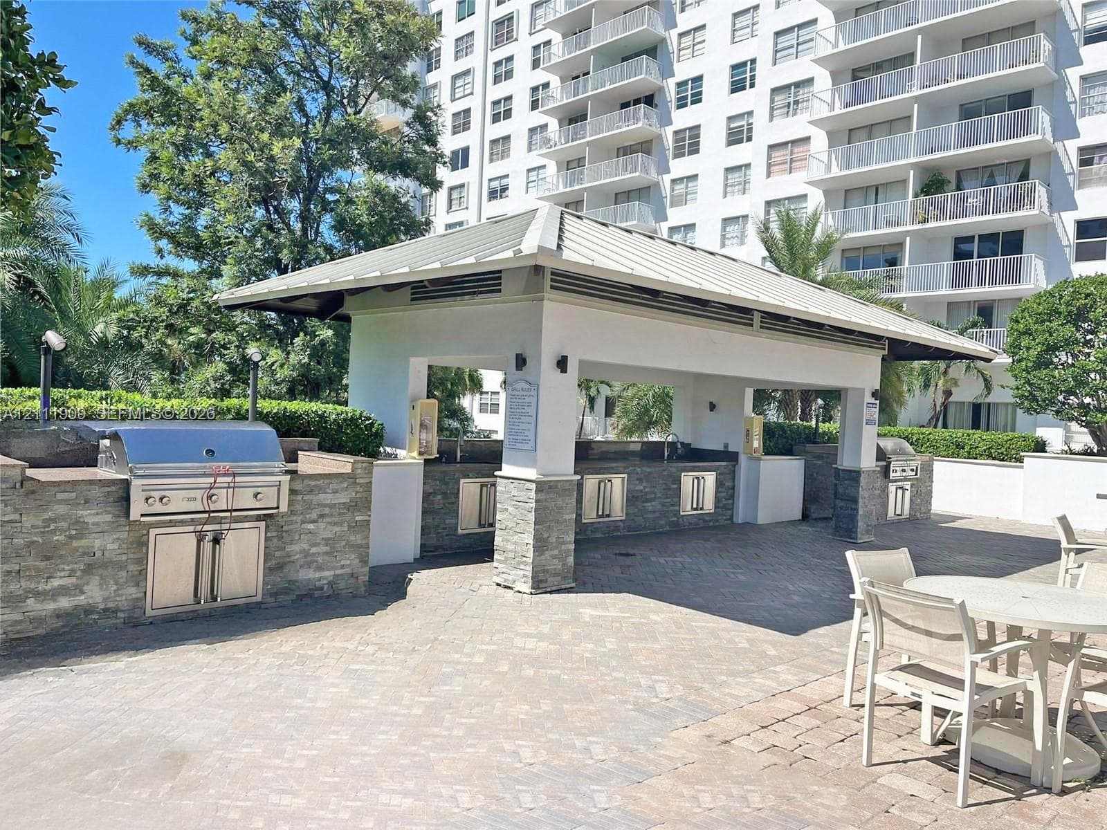 Covered outdoor kitchen featuring stone veneer base cabinetry, built-in stainless steel appliances, a metal roof structure, and brick paver flooring