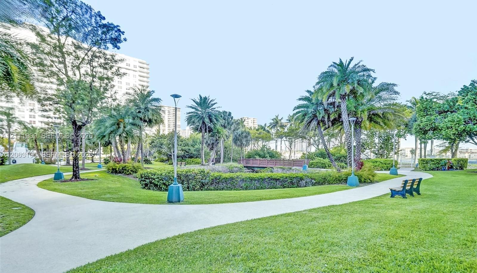 Landscaped grounds featuring winding concrete pathways, mature palm trees, decorative shrubbery, and a wooden pedestrian bridge