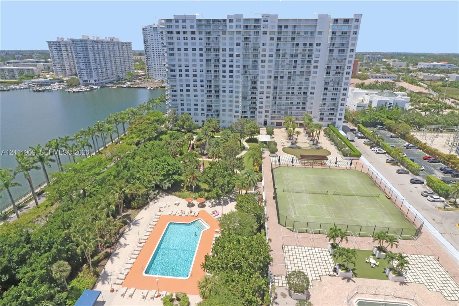 Aerial perspective of a high-rise residential complex featuring a swimming pool, tennis court, landscaped grounds with mature palm trees, and waterfront access