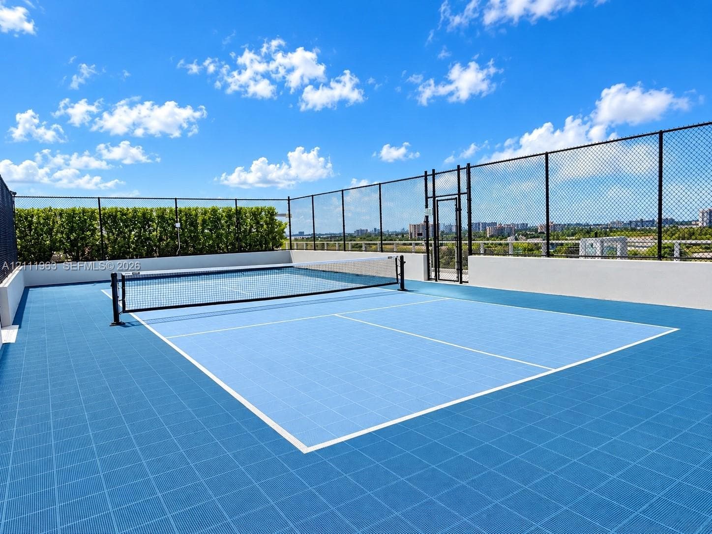 Rooftop tennis court with blue athletic tile surfacing, perimeter chain-link fencing, and integrated privacy hedges