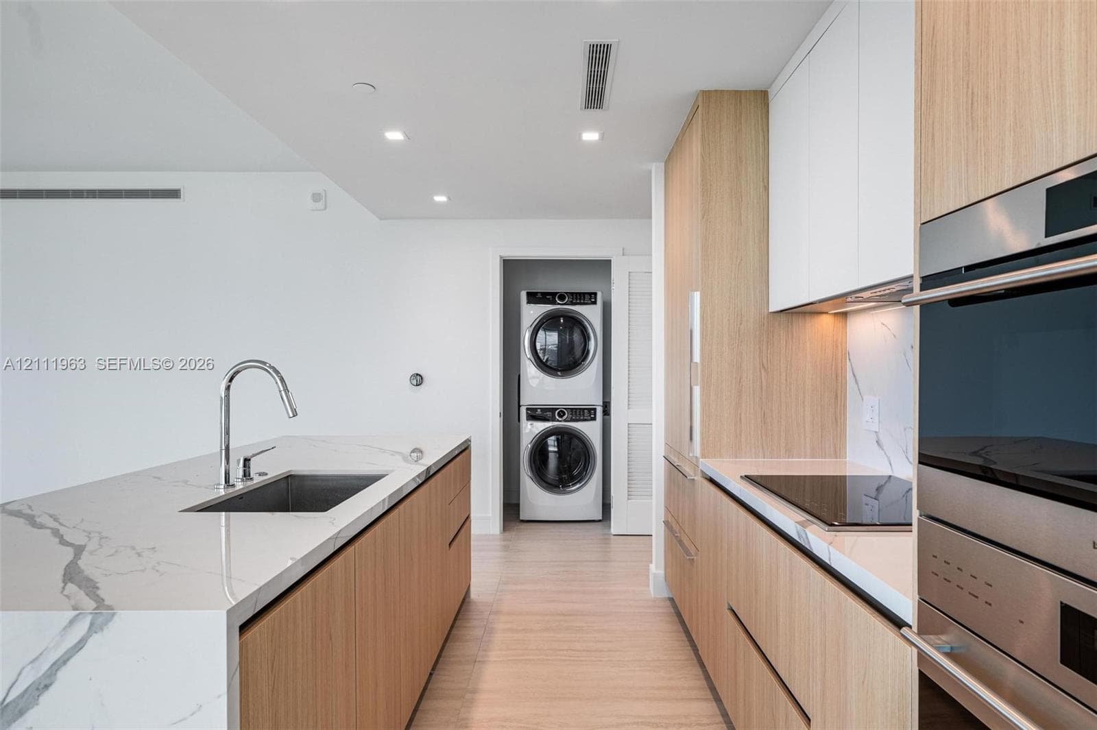 Kitchen featuring light wood-finish cabinetry, waterfall-edge stone island with an undermount sink, integrated stainless steel oven, stone backsplash, and a laundry closet with a stacked washer and dryer