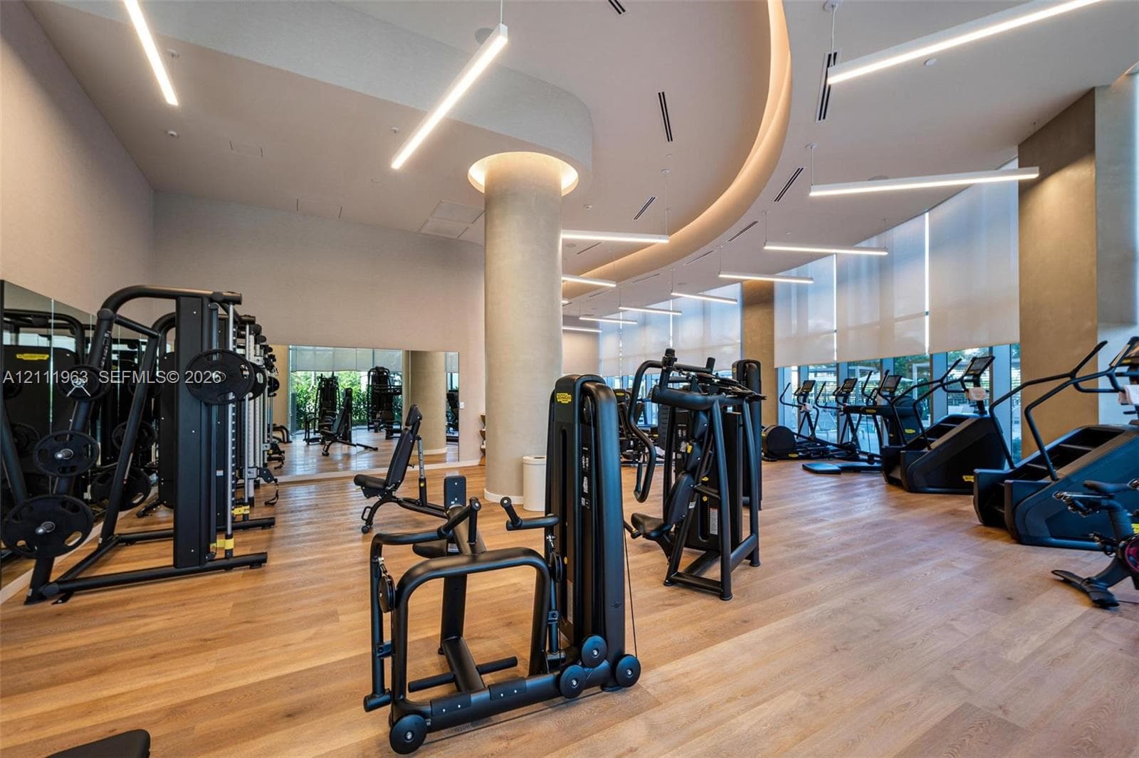 Fitness center featuring wood-finish flooring, floor-to-ceiling windows with roller shades, recessed cove lighting, suspended linear light fixtures, and a structural column