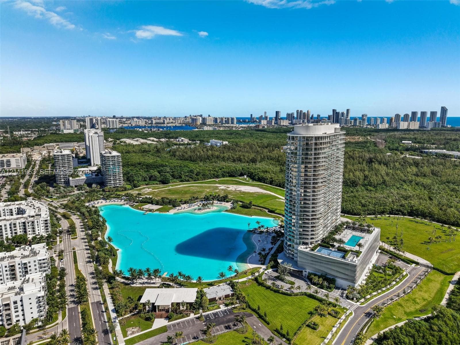 Aerial perspective of a high-rise residential tower featuring an on-site swimming pool, expansive turquoise lagoon, landscaped green spaces, and parking facilities