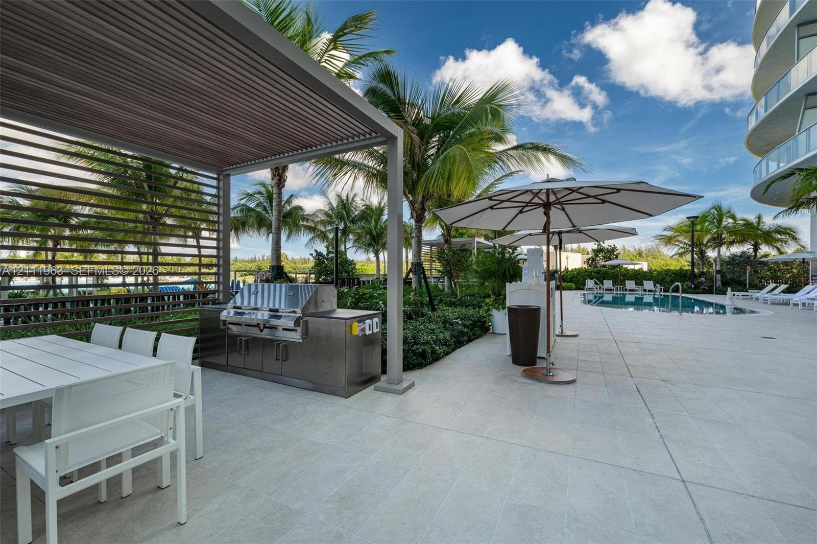Outdoor grilling area featuring a stainless steel built-in barbecue station under a louvered metal pergola, large-format tile patio, and swimming pool
