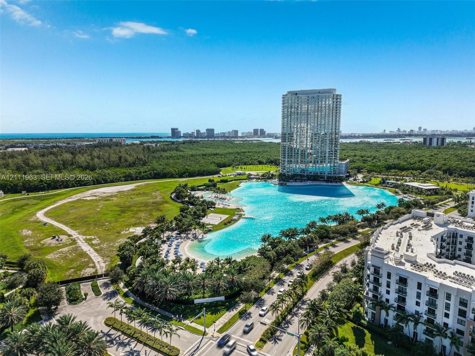 Aerial perspective of a high-rise residential structure featuring a large-scale lagoon, landscaped palm tree surroundings, paved pathways, and an expansive coastal horizon