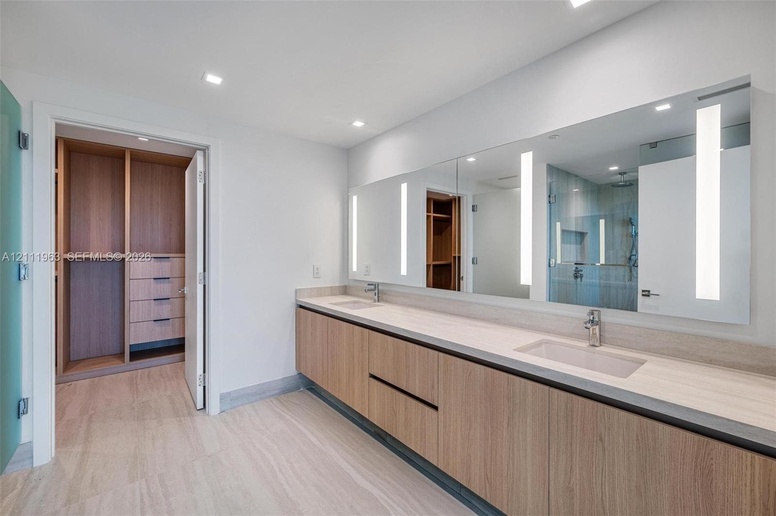 Floating wood-finish vanity with dual integrated sinks, light-toned stone-finish countertop, full-width mirror with integrated vertical LED lighting, and built-in wood cabinetry in the walk-in closet