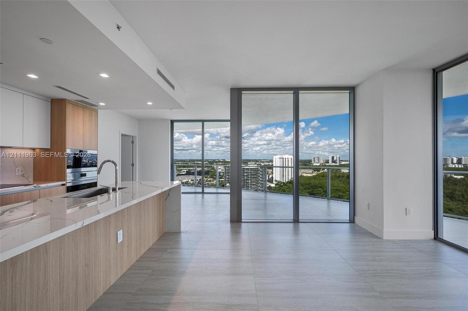 Modern kitchen featuring wood-finish cabinetry, a waterfall-edge stone countertop, built-in stainless steel appliances, floor-to-ceiling glass panel walls, and large-format tile flooring