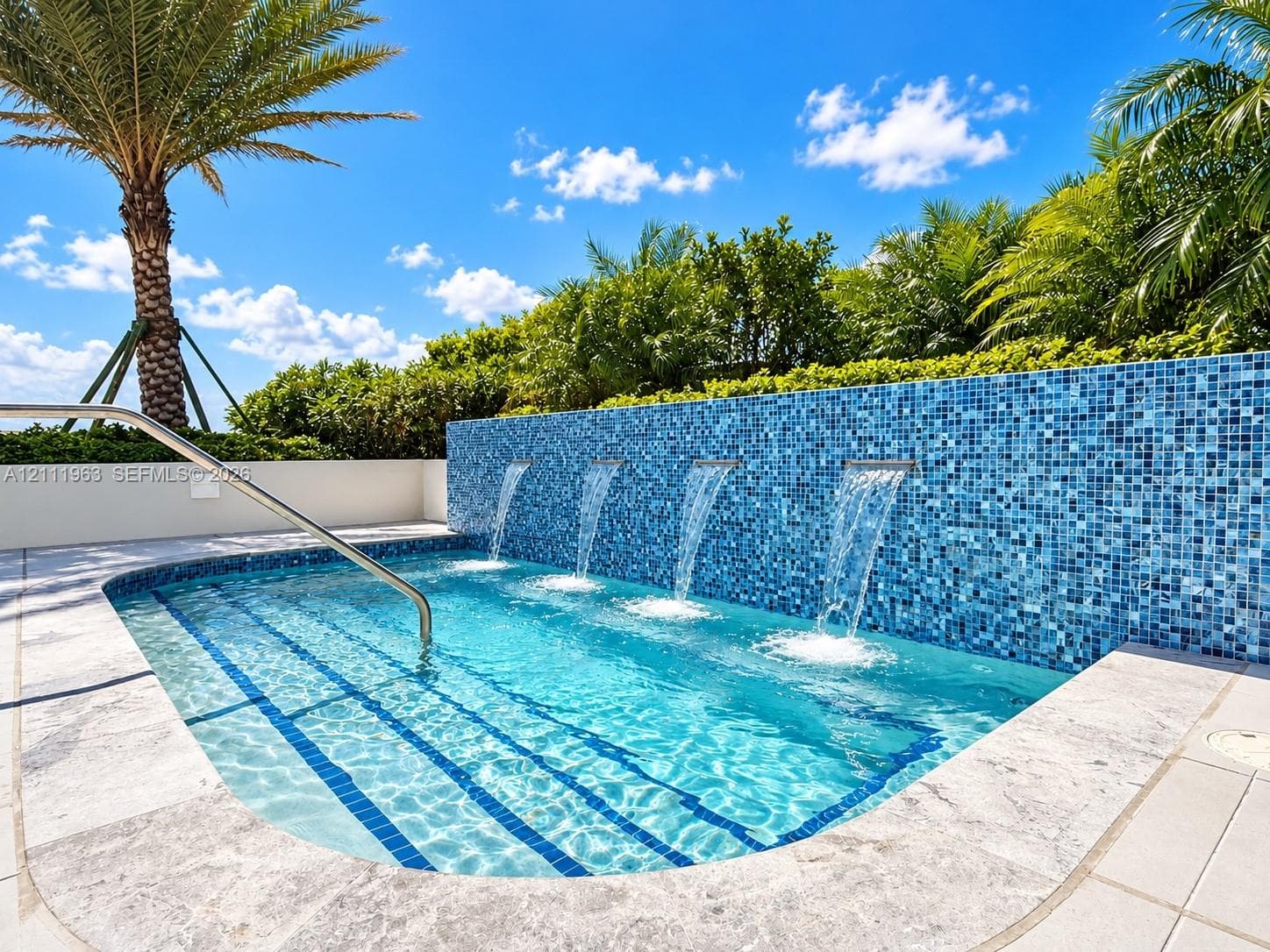 In-ground swimming pool featuring a mosaic tile water feature with four integrated spillways, stone-style pool decking, and a stainless steel entry rail
