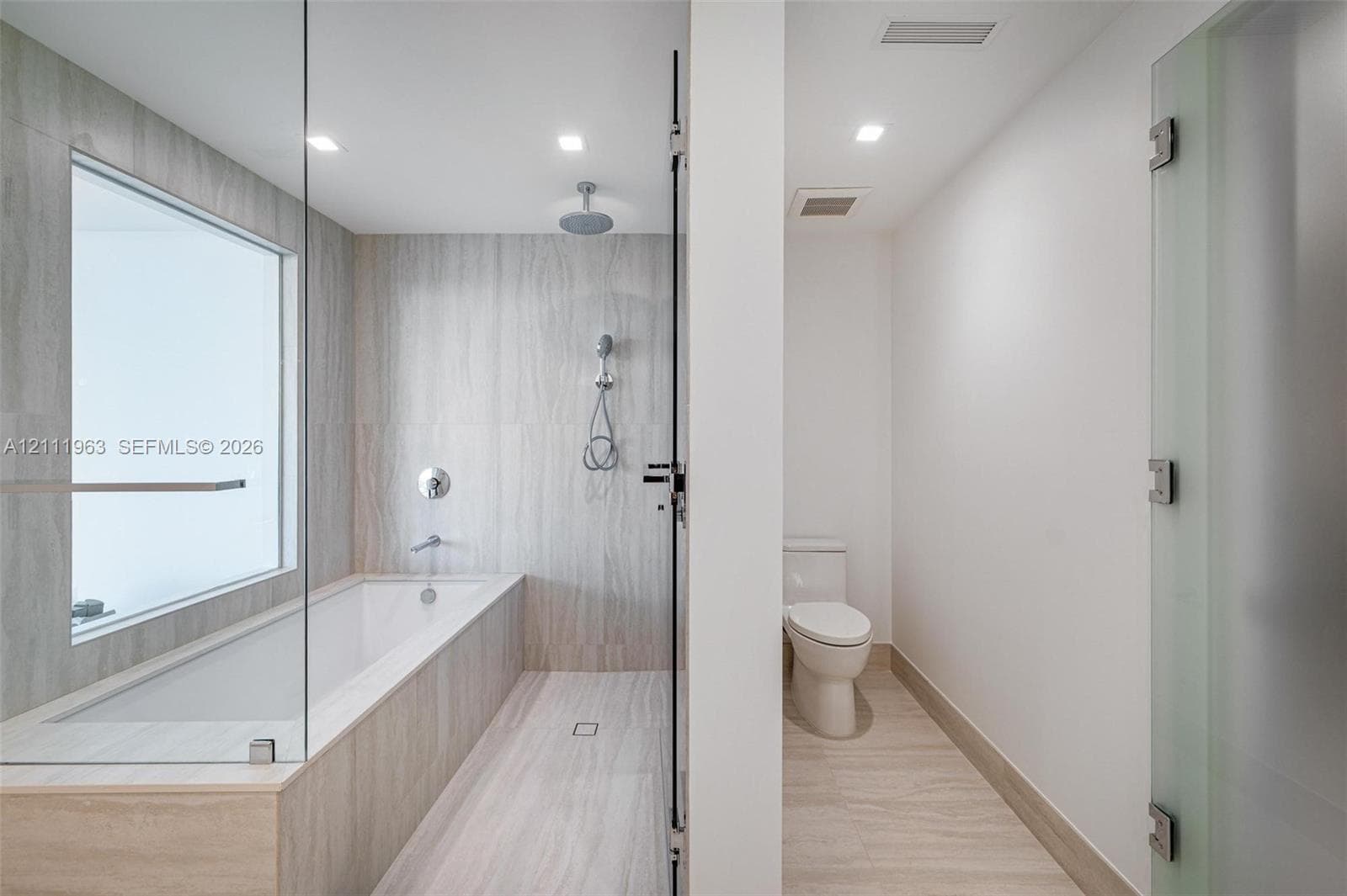 Full-height glass shower enclosure, built-in soaking tub with wall-mounted faucet, rainfall showerhead with handheld sprayer, stone-finish tile surround, and separate enclosed water closet