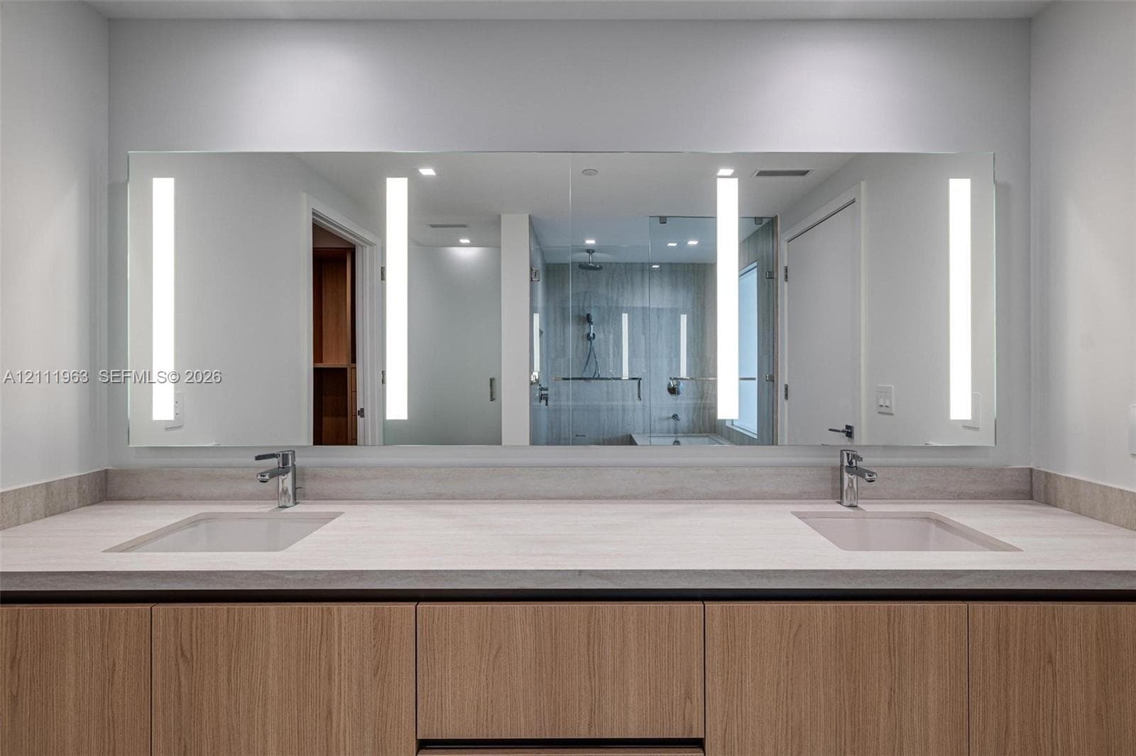 Dual-sink vanity featuring a light-toned countertop, wood-finish cabinetry, integrated rectangular basins, wall-mounted faucets, and an expansive mirror with integrated vertical lighting