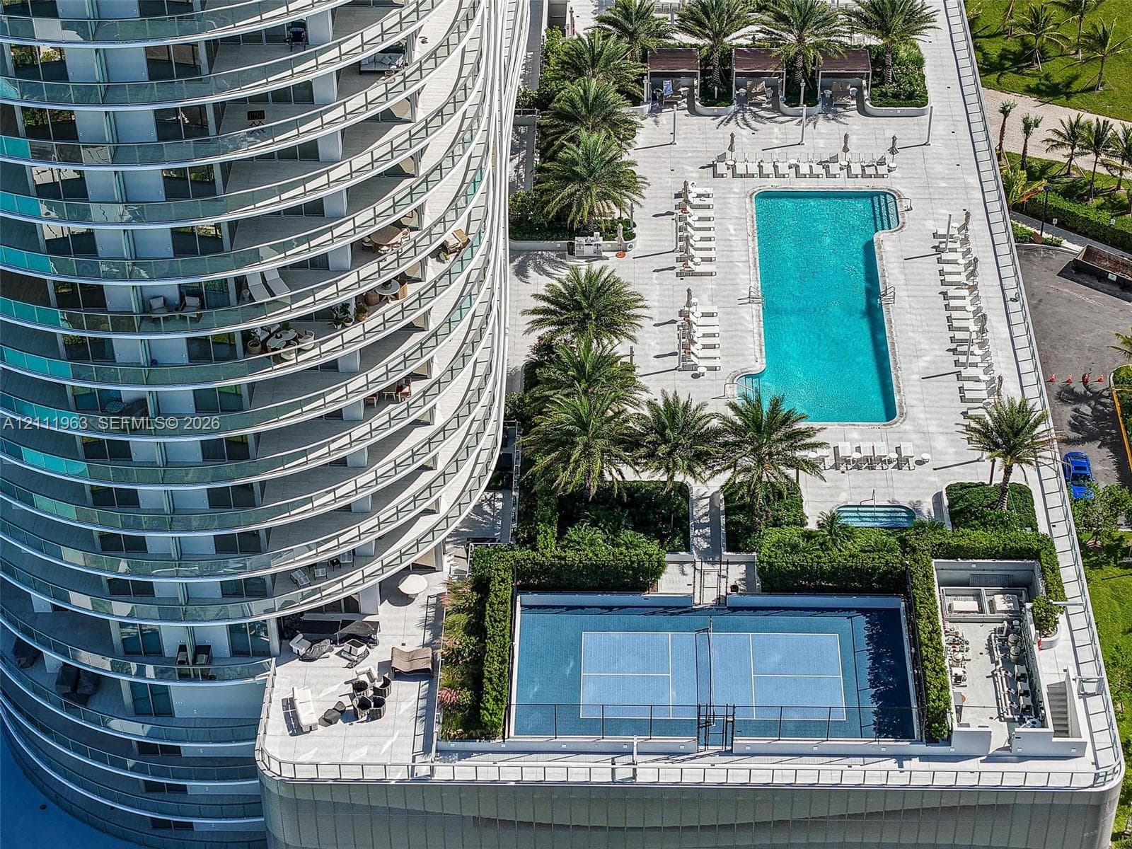 High-rise residential exterior featuring expansive wrap-around balconies, a large swimming pool, a dedicated tennis court, and manicured landscaping with mature palm trees
