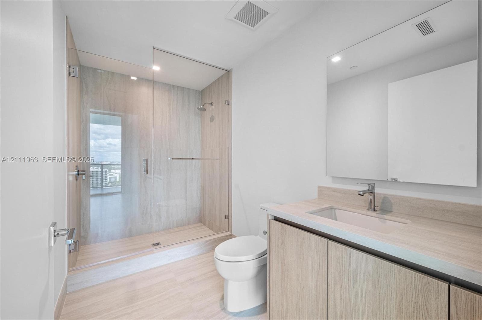 Modern vanity featuring light wood-finish cabinetry and an integrated countertop with a polished chrome faucet, oversized frameless wall mirror, and walk-in shower with floor-to-ceiling tile surround and glass enclosure