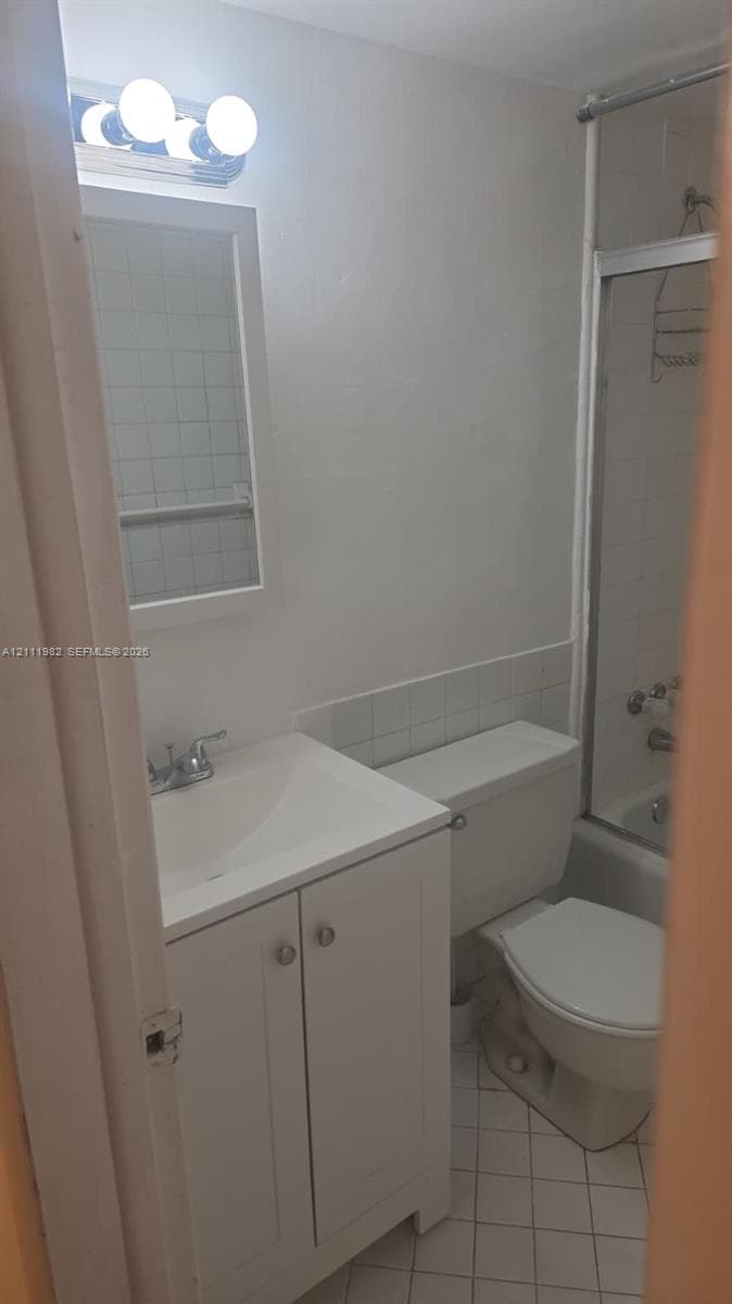 Bathroom featuring a white vanity cabinet, ceramic tile flooring, a shower-tub combination with a glass enclosure, and a vanity-mounted mirror with integrated light fixture
